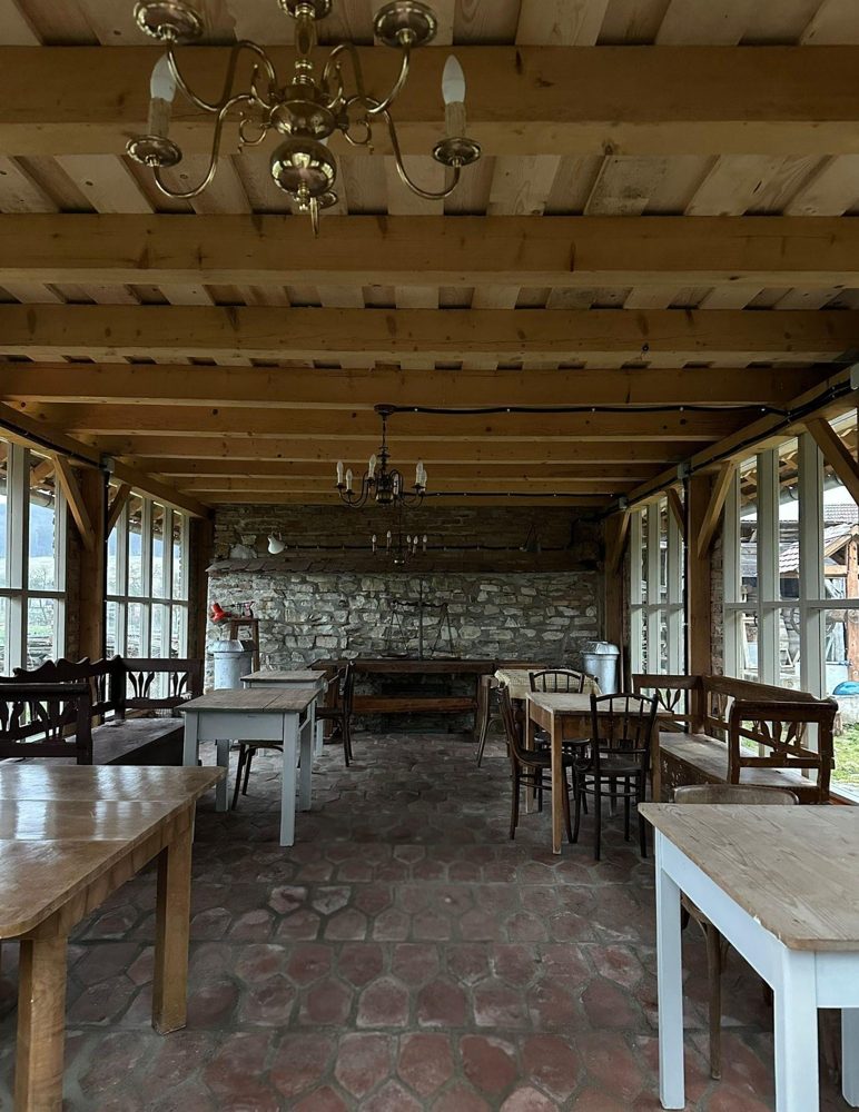 Orangeria — interior cu pereți vitrați, grinzi de lemn masiv, mese și bănci rustice și candelabre din alamă